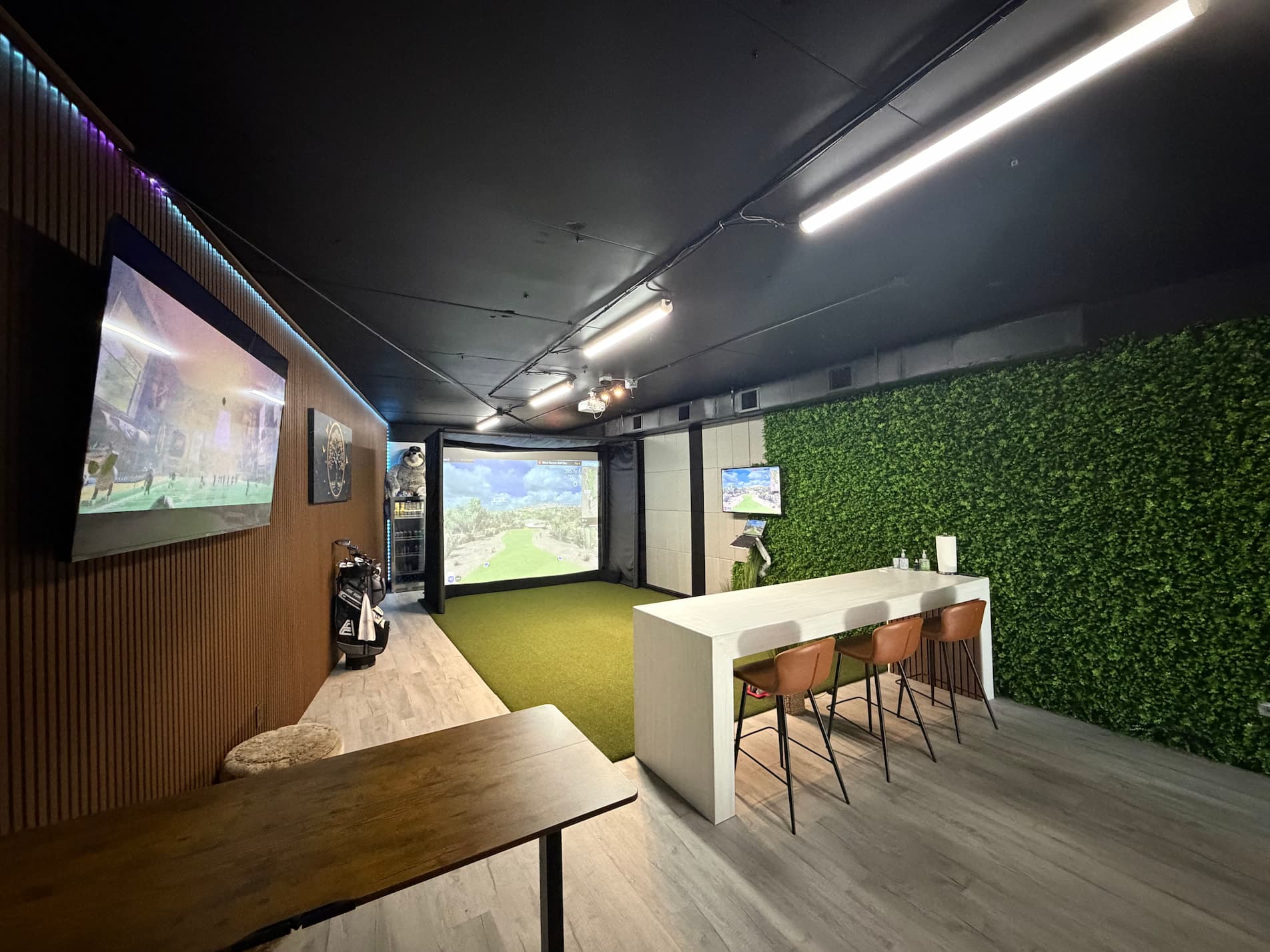 Golf simulator bay with projection screen showing golf course, seating area with bar stools, and green living wall at Golf Lounge Satellite Beach FL